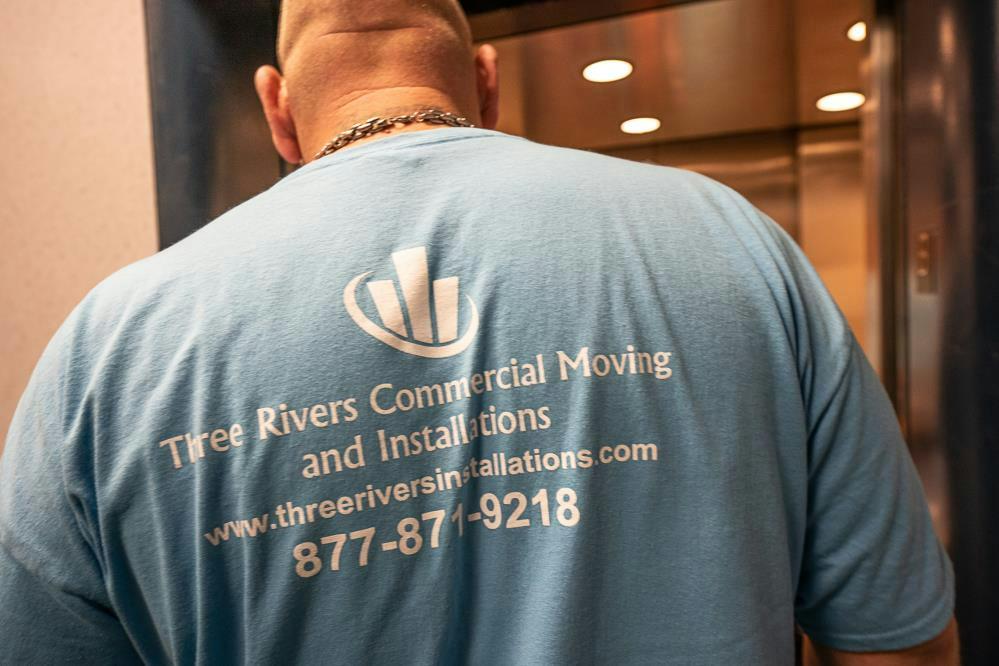Moving guy with his back to the camera sporting a baby blue shirt and white lettering showing the Three Rivers Moving and Installations Logo and phone number